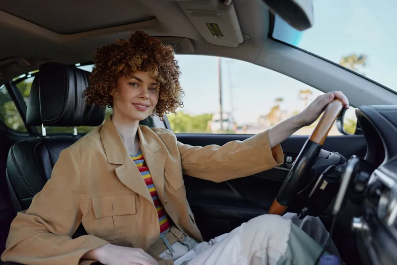 Woman driver sits behind the wheel in a beige jacket and striped shirt, relaxed smile, retro film color palette, daylight through the car window, lifestyle portrait