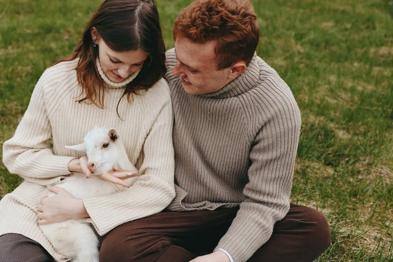 Smiling couple outdoors with a white goat kid, cozy sweaters, gentle touch, and warm light. Tender moment of lifestyle warmth and affection in a grassy meadow with animal companionship.