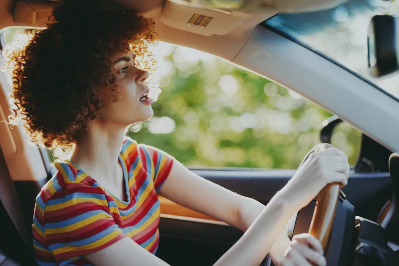Smiling woman driving a car with a striped shirt, looking ahead with focus. Retro film color tones, warm natural light, casual lifestyle moment, everyday travel and independence.