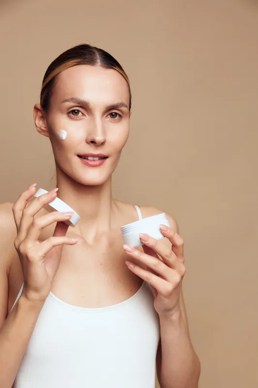 Young woman applies face cream with fingers holds jar of moisturizer looks at camera with soft smile isolated on beige background skincare beauty concept natural glow
