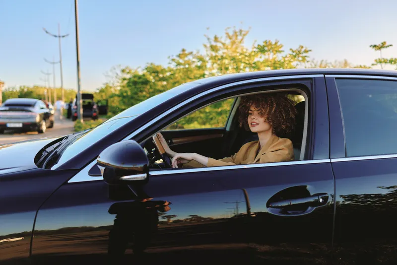 Young woman with curly hair drives a sleek black car, smiling softly as warm sunlight reflects on the window and interior, conveying confidence and everyday motion.
