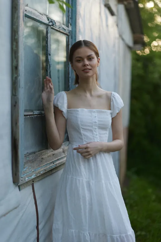 Young woman in a white summer dress standing by a rustic window, exuding a serene emotion in a natural green setting, ideal for seasonal fashion and lifestyle concepts