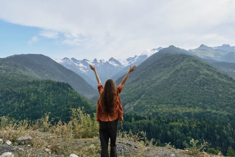 A woman stands on a rocky overlook with arms raised, embracing vast mountains and green valleys in a dramatic nature landscape.
