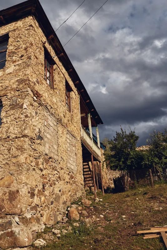 Weathered stone building with wooden balcony and exterior stairs under a cloudy sky, a rural setting with overgrown ground, capturing rustic architectural charm and historic atmosphere.