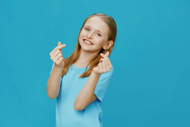 Happy young girl in blue t shirt showing heart gesture on a vibrant blue background, expressing joy and positivity, suitable for family and children oriented content