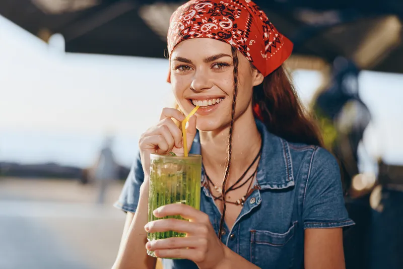 Lifestyle woman in modern boho-western denim street style smiling while drinking a refreshing green smoothie outdoors in warm natural light with film color effect