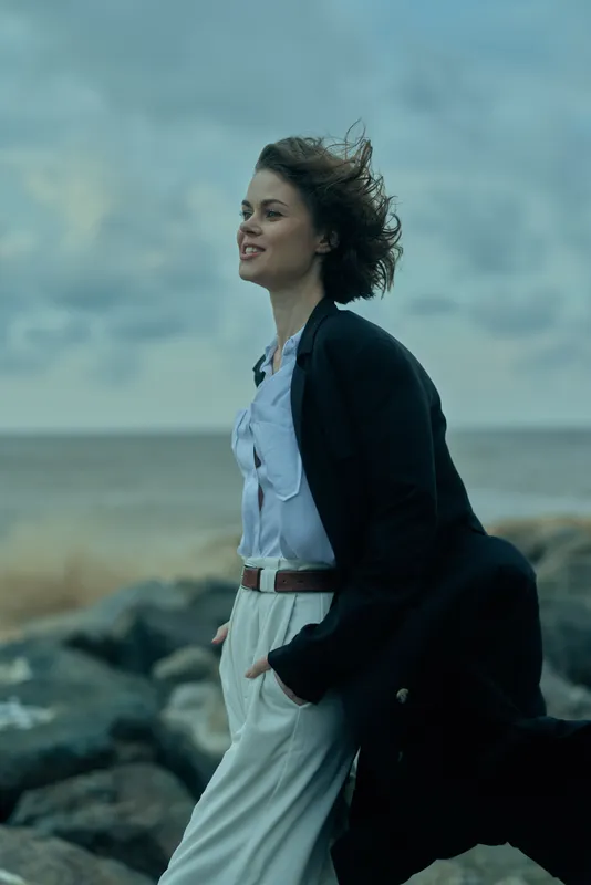 Windy coastal scene with a woman in a flowing dress and a dark coat, standing by rugged rocks as waves roll in, hair swept by wind, capturing motion, style and a fresh ocean atmosphere