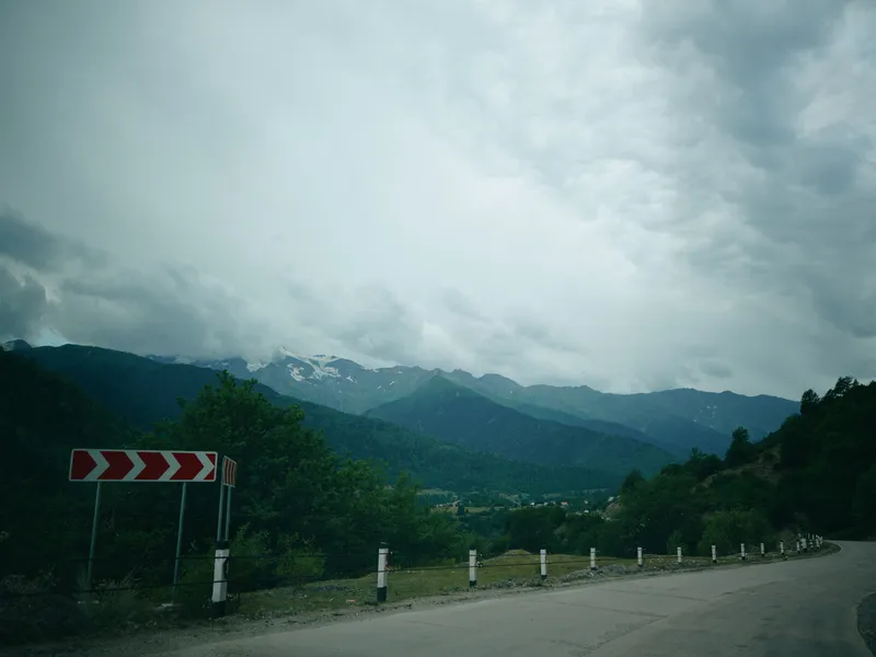 Road landscape with mountains under cloudy sky, guardrail along a winding curve, moody overcast weather creating dramatic travel scenery for nature, outdoors and countryside exploration