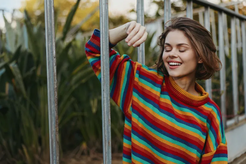 Woman young adult smiling and leaning against a metal railing outdoors in a colorful rainbow sweater, eyes closed, casual outfit, relaxed lifestyle portrait in greenery.