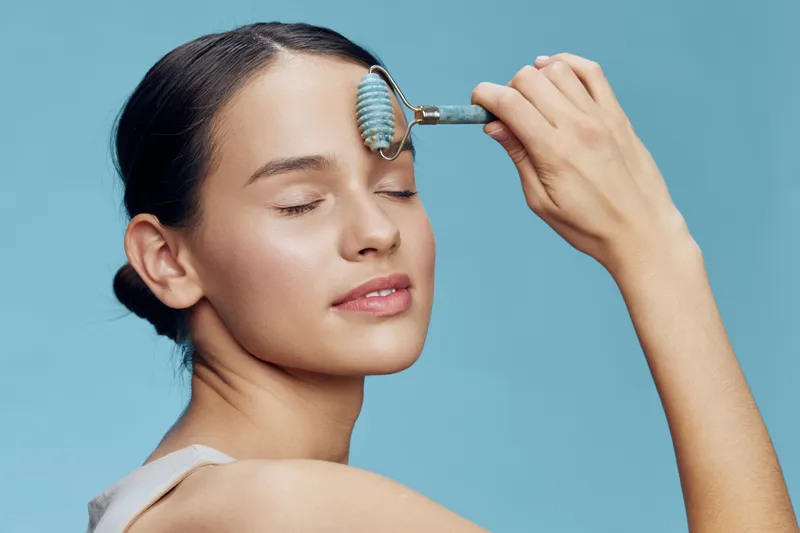 Young woman using a facial roller on her forehead, showcasing skincare routine for healthy skin, serenity, and self care against a light blue background