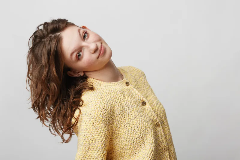 Young woman with curly hair wearing yellow knitted sweater tilts head and smiles gently on white background. Concept of casual style, natural beauty, and relaxed mood.