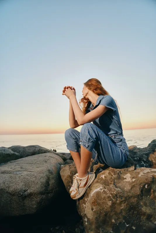 Lifestyle woman in modern boho-western denim street style sitting on rocks at sunset with film color effect, relaxed mood, casual outfit, and natural background.