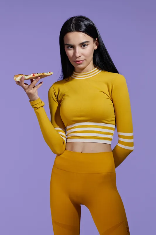 Sport fitness woman holding a slice of pizza while wearing yellow activewear with stripes, confident expression, dark long hair against a purple background.