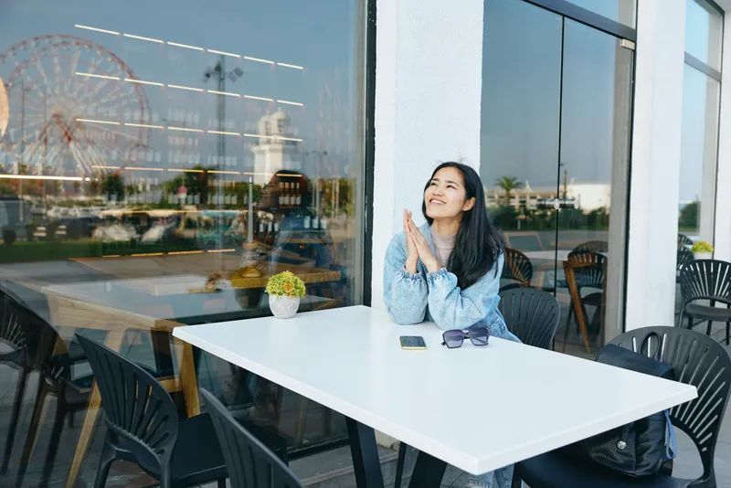 Woman at outdoor cafe table, smiling, denim jacket, glass windows, sunlit urban setting, relaxed mood, casual elegance, table arrangement and chairs in view, reflective ambiance, personal moment