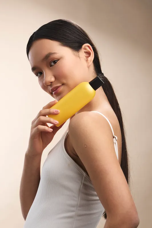 Asian woman in a white top holds a yellow phone to her ear, smiling softly as she chats. The portrait conveys a calm, confident moment with warm lighting and a simple studio backdrop.