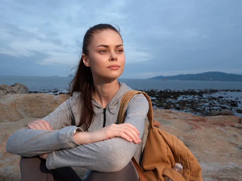 Young woman with a backpack sitting on rocks by the sea, looking thoughtfully at the horizon during a calm summer twilight, embodying peace and contemplation