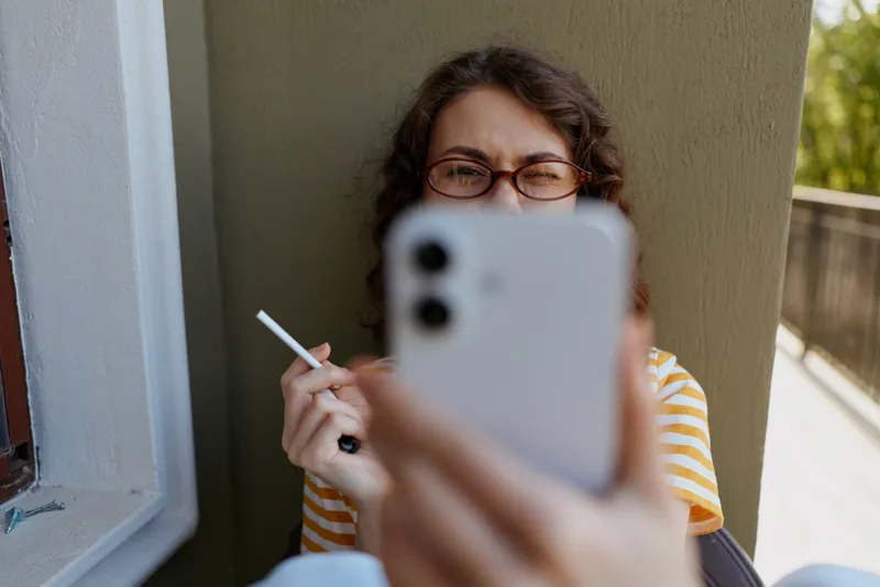 Woman outdoors on a balcony, taking a selfie with smartphone, wearing glasses and striped shirt, relaxed mood, natural light, candid lifestyle moment, soft breeze, greenery in the background.