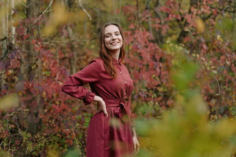 Authentic portrait of a smiling woman in a burgundy dress standing among autumnal shrubs, conveying real life simplicity, warmth, and sincerity in a candid outdoor setting.