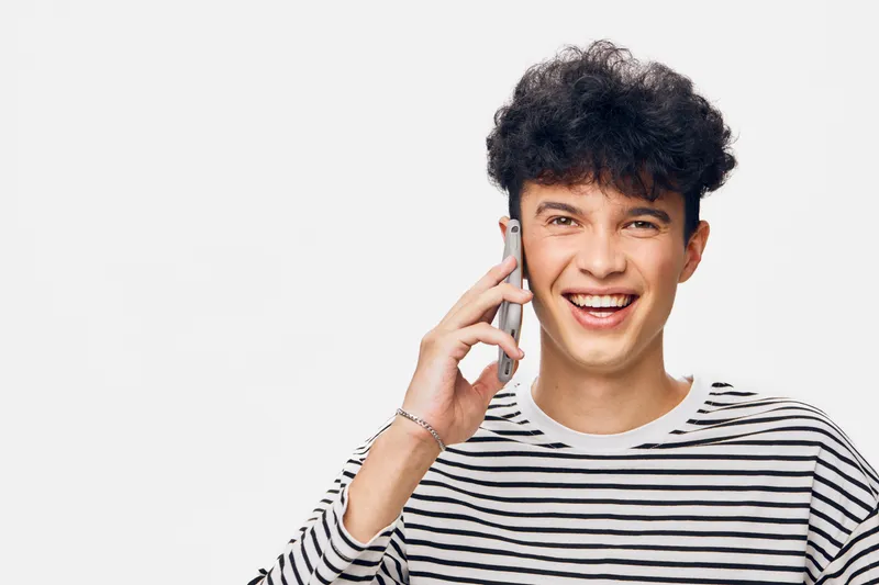 smiling man talking phone in striped shirt, casual young adult with curly hair, cheerful expression on white background, communication concept