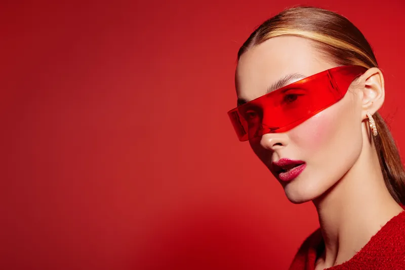 Stylish woman with sleek hair wears futuristic red visor glasses and matching lipstick. Close-up portrait on red background. Concept of fashion, modern style, and bold makeup look.