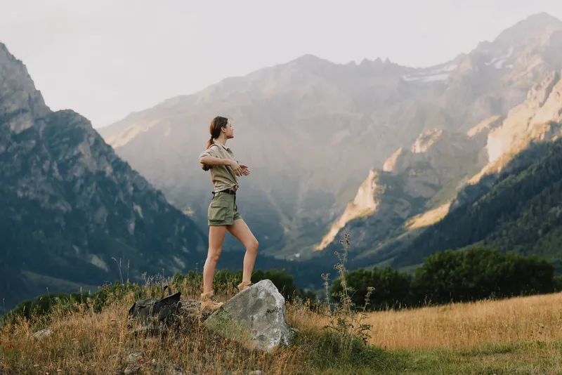 Young woman stands on a rock in an alpine meadow, arms crossed, mountains in the distance, sunlit and tranquil, wildflowers sway softly, clear blue sky.