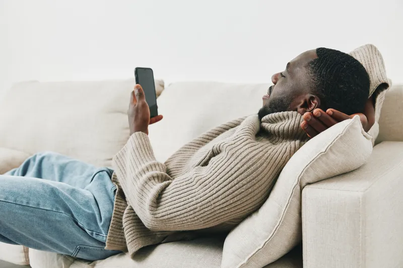Relaxed man of African descent lounging on a sofa while using a smartphone, showcasing comfort, casual wear, and modern lifestyle