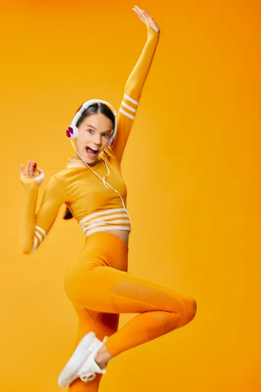 Vibrant image of a woman fitness enthusiast in orange athletic wear jumping with headphones, exuding energy and motivation in a bright studio backdrop.