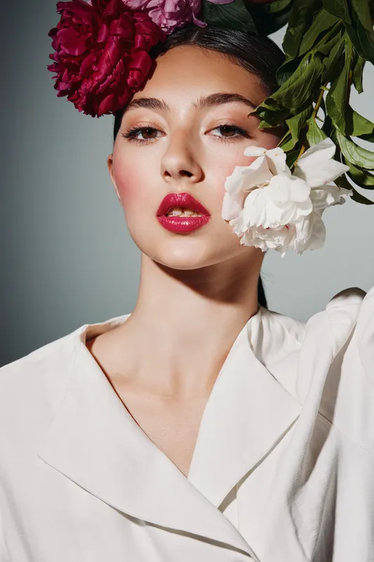 Fashion model with vibrant red and white floral headpiece poses wearing bold makeup and elegant white garment, showcasing creative beauty styling in studio light. Concept of fashion and beauty.