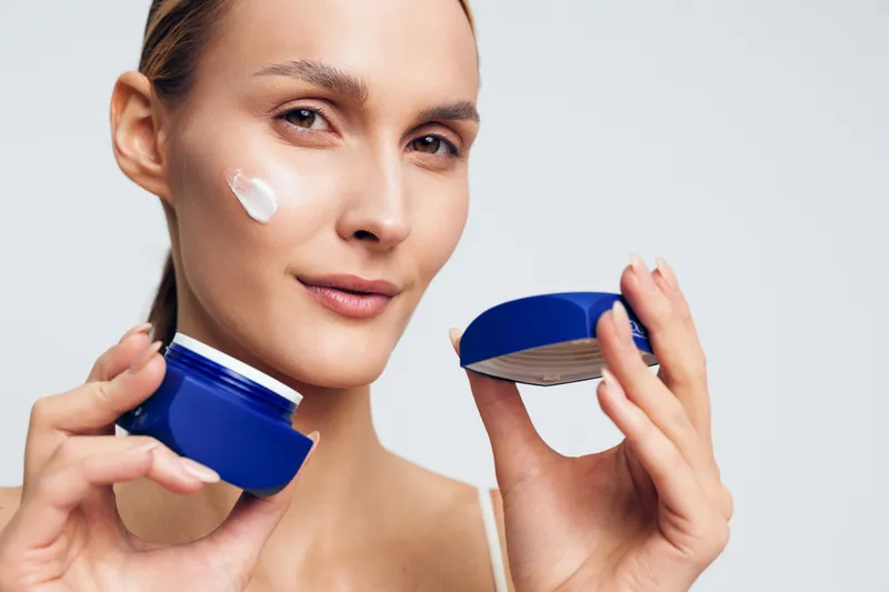 Young woman applies moisturizing cream on face holds blue jar skincare product closeup isolated on white background natural beauty care concept with smooth skin and gentle expression