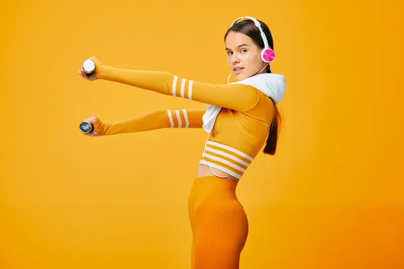 A woman fitness concept with sport vibe: an athletic woman in orange outfit wears headphones and stretches arms while holding small dumbbells in a bright orange studio, conveying energy, balance and