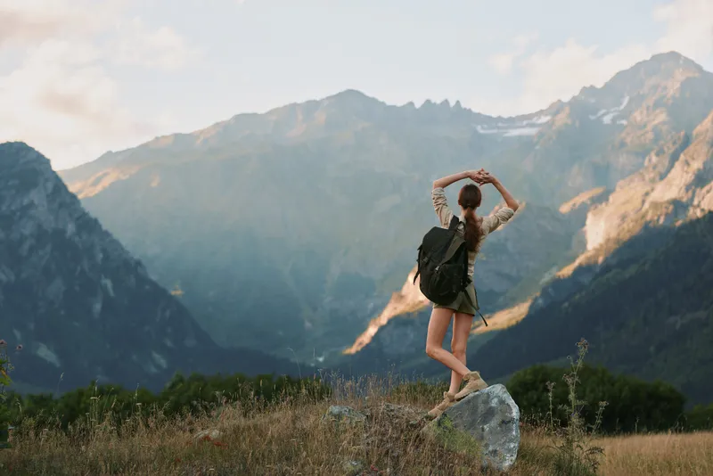 Woman hiking in the mountains with a backpack, standing on a rock. Outdoor scene highlighting nature, adventure, and personal exploration in a broad alpine landscape.