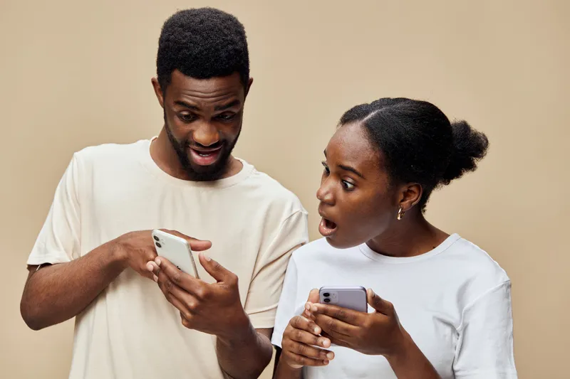 Young couple surprised by something on their smartphones, expressing shock and intrigue against a warm background, ideal for technology and communication themes