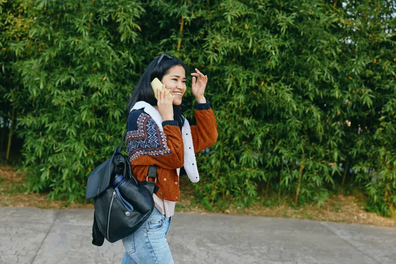 Teenager girl talking on two phones outdoors, smiling, with backpack and casual jacket, beside green hedge and concrete path, capturing contemporary street style and multitasking moment