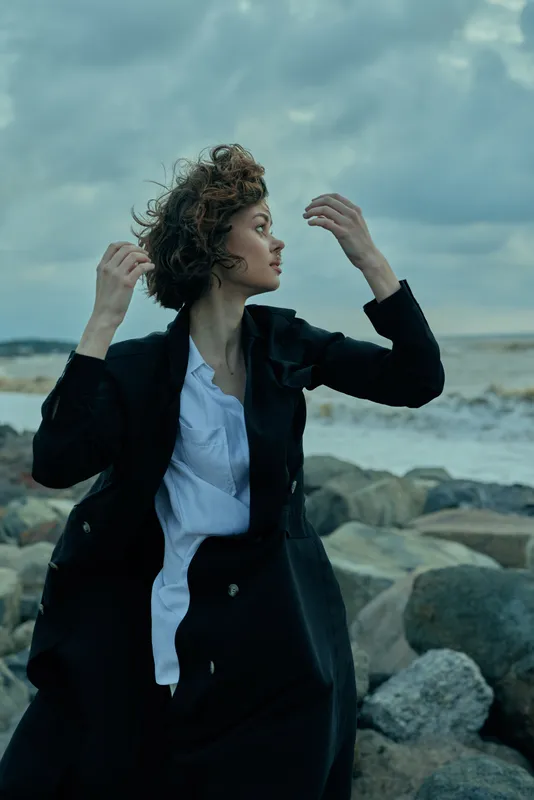 Woman in a black coat and white shirt on a windy coast with rocks, expressing movement and fashion in an outdoor sea scene