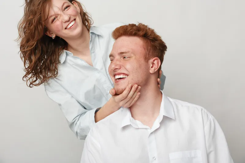 Happy young couple with red hair and casual white shirts smiling and enjoying playful moment together on light background. Concept of love, friendship, joy, and positive emotions.