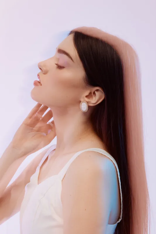 Young woman with long brown hair wearing white earrings and a white top poses with eyes closed and hand near neck. Soft lighting highlights her beauty and fashion style. Studio shot.