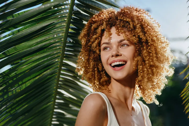 smiling woman with curly hair enjoying sunny tropical outdoors, natural light, green palm leaves, carefree mood, summer vibes