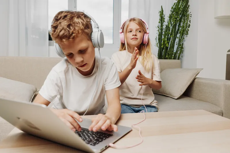 Two children with headphones enjoy technology at home, a boy focused on laptop and a girl listening to music, sitting on a sofa with casual clothes and natural light.
