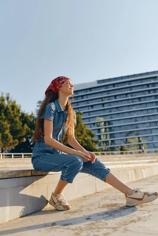 young woman in casual denim outfit sitting outdoors with red headscarf, enjoying sunny day by modern building and green trees, relaxed thoughtful mood