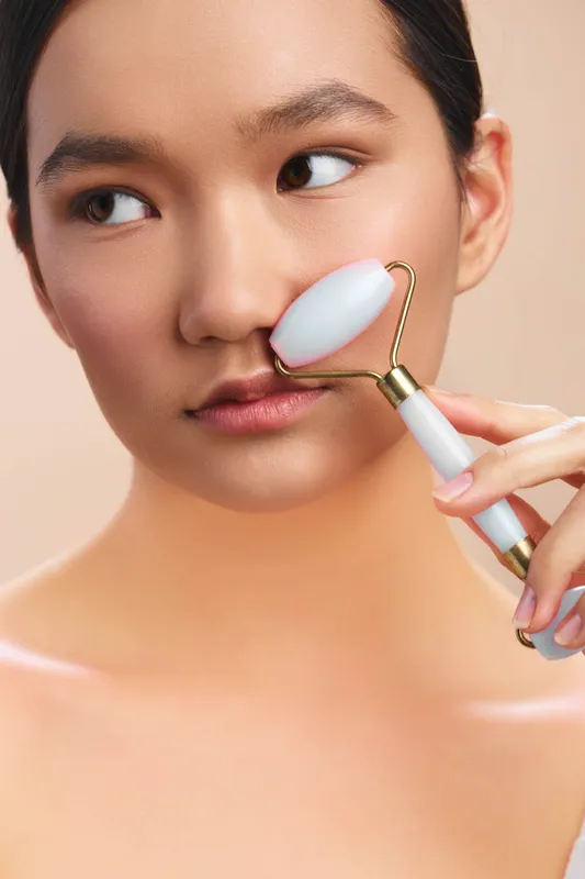 Asian woman performs a facial rolling routine with a gentle expression, using a soothing skincare tool on her cheek in a serene, closeup portrait with a calm mood.