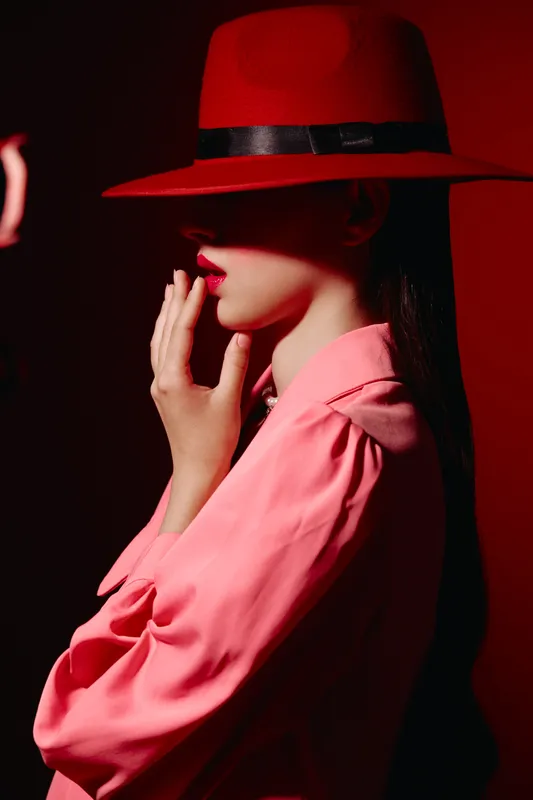 Stylish woman in a vivid red hat and pink blouse poses gracefully in low light. Emphasis on fashion, beauty, and creative portrait with dramatic shadows and smooth skin tones.