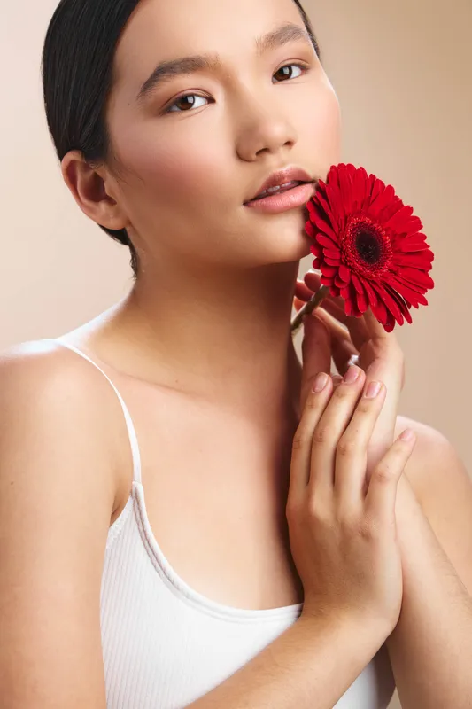 Asian woman gracefully holds a vibrant red flower near her face, conveying elegance and calm. Soft lighting highlights smooth skin and a serene, confident expression.