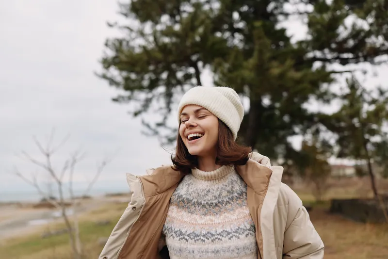 Woman smiling in a cozy winter outfit stands outdoors among trees, wearing a warm jacket, sweater and beanie, capturing a joyful portrait amid cool natural light and serene countryside