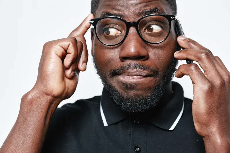 African American man with dark skin wearing glasses and black polo shirt talking on smartphone and thinking with finger on temple isolated on white background.