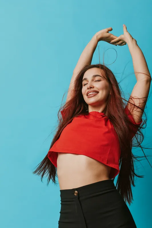 Young woman with long hair wearing red crop top and black pants stretching arms up with eyes closed and smiling on blue background. Relaxation, happiness and freedom concept.
