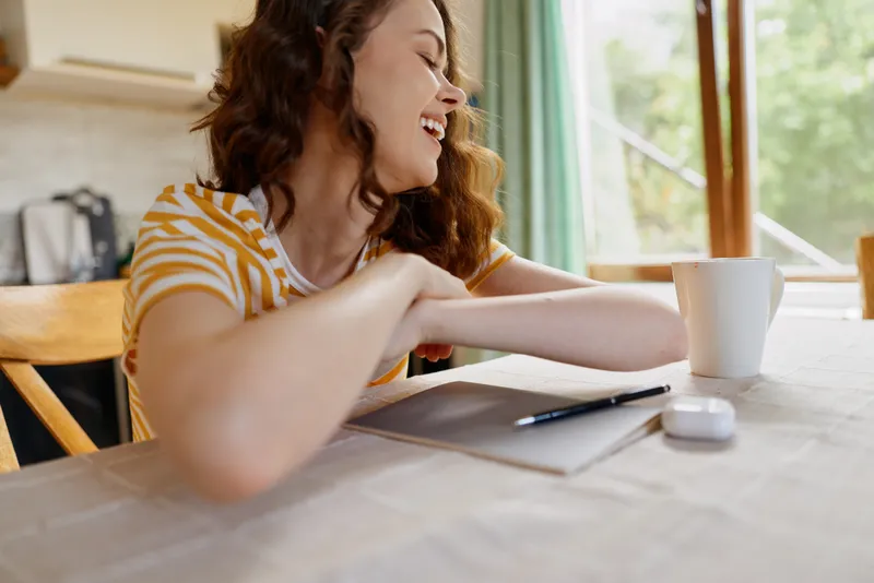 Smiling woman with curly hair seated at a bright kitchen table, resting arms on a notebook. Casual home lifestyle, expressing comfort, focus, and creativity.