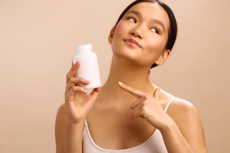 asian woman holds a white bottle and smiles softly, presenting a clean skincare product against a warm neutral backdrop, inviting attention to health and beauty in everyday routines.