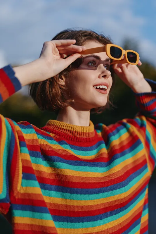 Woman, young woman, striped sweater, sunglasses, smiling, outdoor, portrait, short hair — Young woman in a colorful striped knit and yellow sunglasses looking up and smiling in a sunlit park, casual