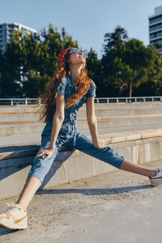 Lifestyle woman in modern boho-western denim street style wearing sunglasses and bandana, sitting outdoors on sunny day with film color effect and casual urban vibe.