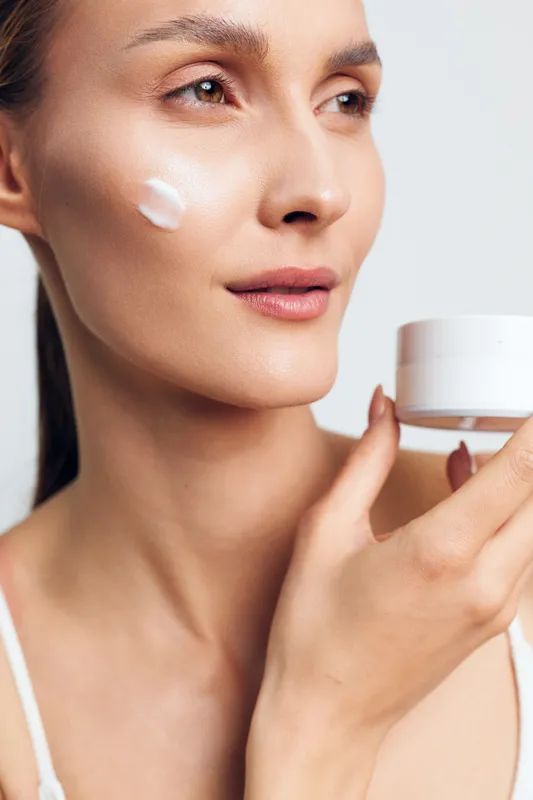 A young woman applies moisturizing cream on her cheek holding a white jar. The close-up shows smooth skin and a natural makeup look in soft lighting. Skincare concept.
